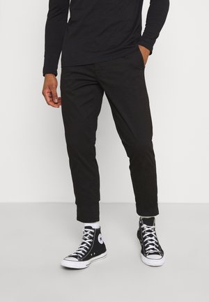 Person trägt schwarze Slim-Fit-Hose, schwarze High-Top-Sneaker mit weißen Schnürsenkeln und ein schwarzes Langarmshirt und steht mit einer Hand in der Tasche.