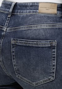 Poche arrière d'un jean en denim bleu avec des coutures visibles et une étiquette beige portant la mention "ANNO 1995 / BLUE DENIM ONLY" sur la ceinture.