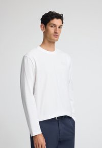 Vit långärmad t-shirt i bomull med rund halsringning och en liten präglad logotyp på bröstet, kombinerad med marinblå byxor.