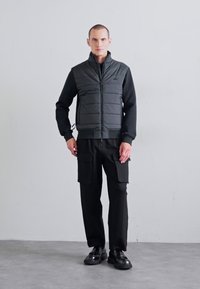 Black, insulated vest with a high collar and zip closure, worn over a long-sleeve black shirt, paired with black cargo pants and shoes.
