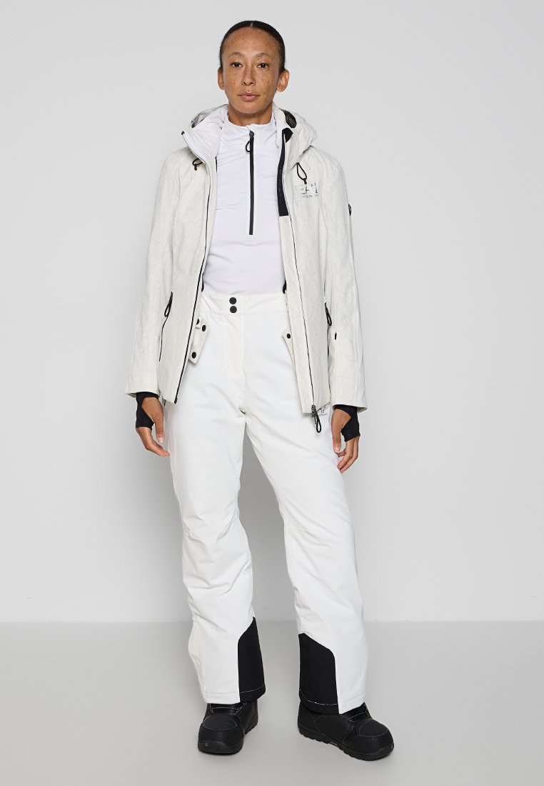 Giacca impermeabile bianca con cappuccio e zip, abbinata a pantaloni da neve bianchi con dettagli neri e bottoni. Stivali neri completano l'outfit.