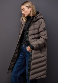 Dlouhý, lehce polstrovaný kabát v taupe s černým zipem a kapucí. Disponuje texturovanou tkaninou, elastickými stahovacími šňůrkami a bočními kapsami. Nositelný přes tmavé džíny.