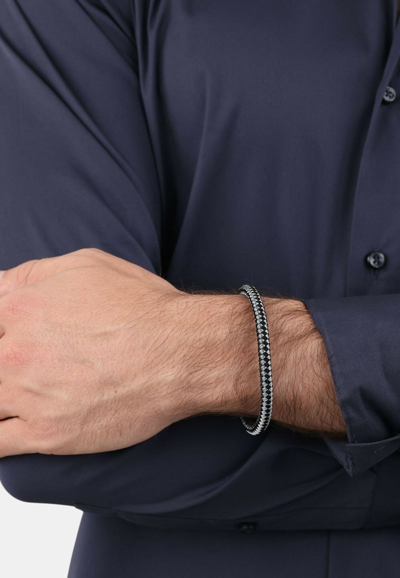Bracciale nero texturizzato con design intrecciato, posato su un polso, abbinato a una camicia a maniche lunghe blu scuro.