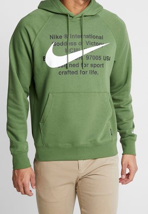 Mężczyzna w zielonej bluzie z kapturem Nike z białym logo Swoosh i czarnym napisem, połączony z beżowymi spodniami, stojący na jednolitym tle.