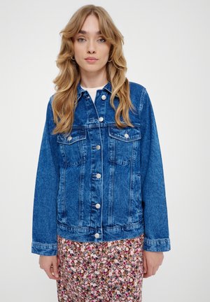 Chaqueta de mezclilla azul con mangas largas, bolsillos frontales y botones plateados; llevada sobre una falda con estampado floral con pequeñas flores de colores.