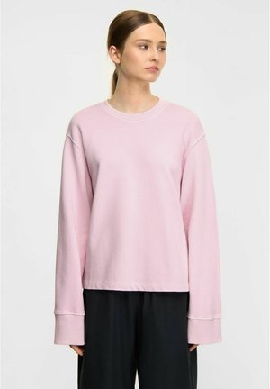 Ung kvinde med tilbagebundet hår iført en løs lyserød langærmet skjorte og sorte bukser, stående foran en ensfarvet baggrund.