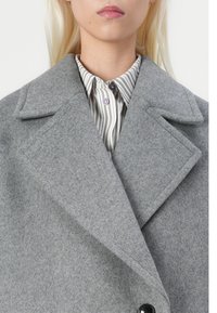 Cappotto grigio in lana con revers a punta, con un unico bottone nero, indossato sopra una camicia a righe con colletto abbottonato.