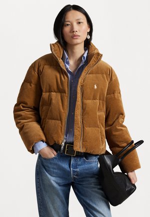 Doudoune en velours côtelé marron avec fermeture éclair, présentant des sections matelassées, associée à une chemise bleu clair et un sac à main noir.
