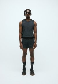 Schwarzes athletisches ärmelloses Oberteil und Shorts aus leichtem Stoff. Zubehör umfasst Sonnenbrille und schwarze Sportschuhe mit strukturierten Sohlen.