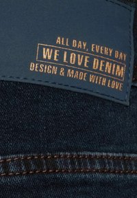 Tissu en denim foncé avec un patch en cuir bleu embossé de texte doré indiquant « ALL DAY, EVERY DAY WE LOVE DENIM DESIGN & MADE WITH LOVE. »