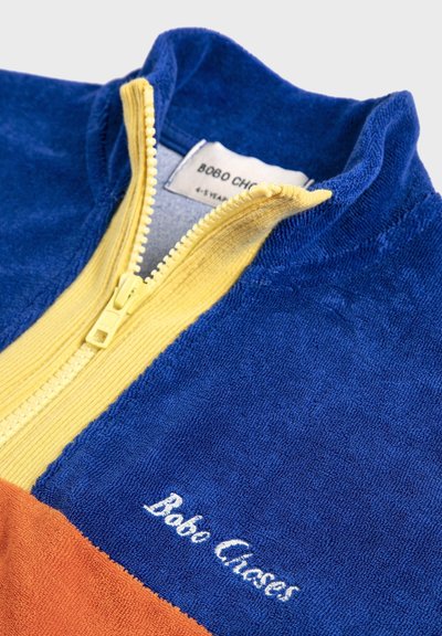 Veste en éponge bleue avec une section inférieure orange, une fermeture éclair jaune partiellement ouverte et "Bobo Closes" brodé sur la poitrine.