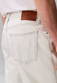 Κοντινό πλάνο ατόμου που φοράει ανοιχτόμπεζ τζιν με καφέ δερμάτινο ταμπελάκι Calvin Klein πάνω από την πίσω τσέπη και λευκό πουκάμισο.