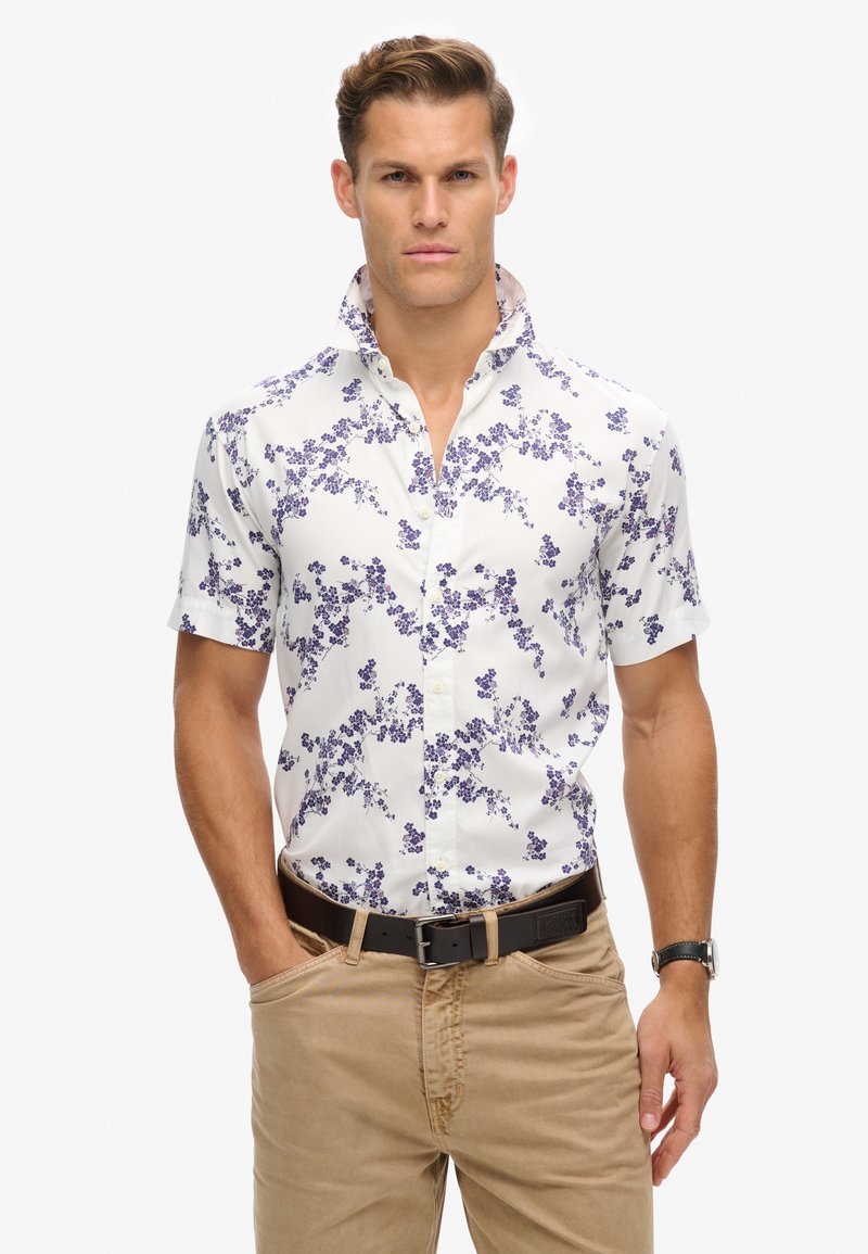 Camisa de manga corta abotonada en blanco con patrón floral púrpura. Cuenta con un cuello tipo solapa y cierre de botones estándar. Diseño casual.