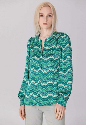 Femme aux cheveux blonds portant un chemisier à manches longues à motif zigzag vert, bleu et blanc et un pantalon gris clair, debout devant un fond uni.