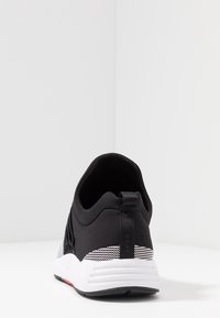 Musta-valkoinen urheilukenkä näkyy takaa, jossa on vetonauha ja teksturoitu valkoinen pohja yksinkertaista valkoista taustaa vasten.