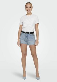 T-shirt bianca con design a farfalla, pantaloni shorts in denim blu, cintura nera e tacchi blu chiaro, il tutto su uno sfondo neutro.