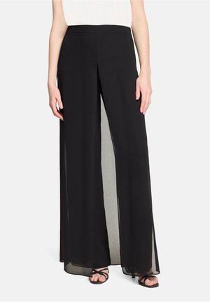 Femme portant un pantalon noir à jambes larges avec des fentes à l'avant, associé à des sandales noires à lanières et un haut blanc, debout devant un fond uni.