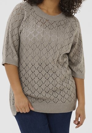Suéter de punto beige con patrón calado en forma de diamante, usado por una persona con cabello rizado oscuro y vaqueros azules, de pie contra un fondo blanco.