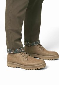 Tans suede enkellaarzen met ronde neus, vetersluiting en gestructureerde rubberen zool. De broek heeft een geruite zoom in donkergroen en marineblauw.