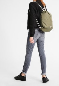 Personne portant une veste noire, un jeans gris, des baskets Reebok noires et portant un sac à dos à cordon olive, se tenant devant un fond blanc.
