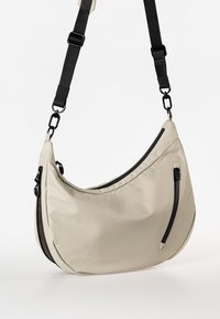 Beige Schultertasche mit geschwungenem Design, aus glattem Stoff gefertigt. Verfügt über eine schwarze Reißverschlusstasche und einen verstellbaren schwarzen Tragegurt.