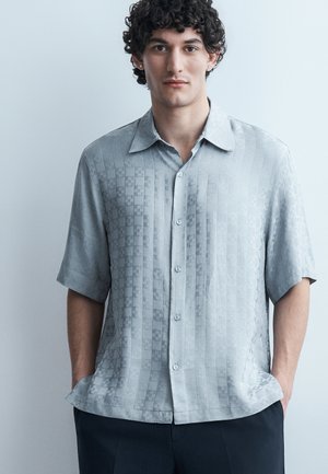 Młody mężczyzna z kręconymi włosami nosi jasnoszarą koszulę z krótkim rękawem i subtelnym kratkowanym wzorem, ręce w kieszeniach ciemnych spodni.