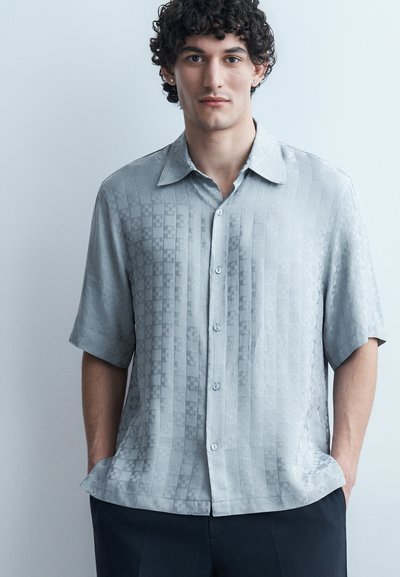 Jeune homme aux cheveux bouclés portant une chemise à manches courtes gris clair à boutons avec un motif à carreaux subtil, les mains dans les poches de son pantalon foncé.