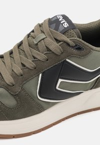 Baskets vert olive en daim et mesh, avec un grand logo noir, des rayures blanches, et une semelle en caoutchouc à motif antidérapant.