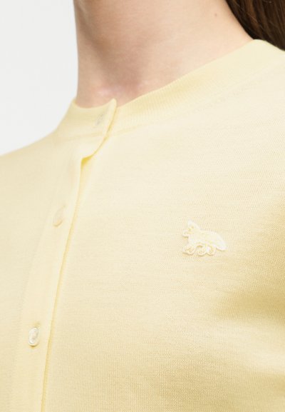 Φοράει ανοιχτό κίτρινο ζακέτα με κουμπιά, που έχει ένα μικρό κεντημένο λογότυπο ζώου στο αριστερό στήθος σε απλό φόντο.