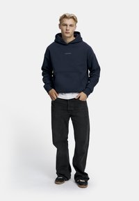 Marineblå hettegenser med frontlomme, hvitt logo, brukt over en hvit skjorte; kombinert med mørke jeans og sorte joggesko. Rett passform.
