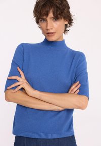 Niebieski sweter z krótkim rękawem, o prążkowym, półwysokim dekolcie, miękkiej teksturze i luźnym kroju, z poziomym prążkowanym wykończeniem u dołu.