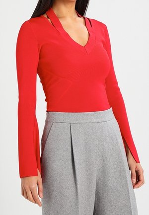 Frau trägt ein rotes Langarmshirt mit Ausschnitt am Hals und graue, weit geschnittene, hoch taillierte Hosen vor weißem Hintergrund.