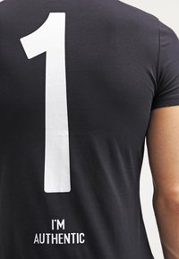 Mustahiili musta t-paita lyhyillä hihoilla, jossa on iso valkoinen "1" selkäpuolella ja teksti "Olen aito" sen alapuolella. Sujuva puuvillakangas.