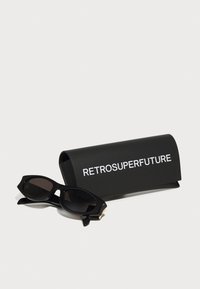Occhiali da sole neri con design angolare, lenti scure e dettagli con logo, accanto a un astuccio nero opaco etichettato "RETROSUPERFUTURE."