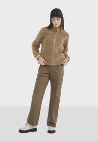 Braune Fleece-Jacke mit Reißverschluss und gerippten Bündchen, kombiniert mit braunen Cargo-Hosen mit seitlichen Taschen. Getragen mit einer schwarzen Mütze und grauen Stiefeln.