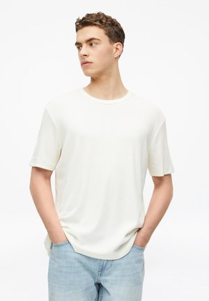 Junger Mann trägt ein schlichtes weißes kurzärmeliges T-Shirt und hellblaue Jeans, steht mit den Händen in den Taschen und schaut nach links.