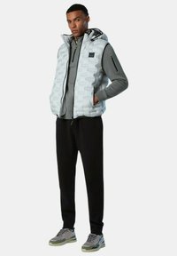 Helles graues gepolstertes West mit Kapuze, Reißverschluss und strukturiertem Muster, getragen über einem grauen Langarmoberteil, kombiniert mit schwarzen Hosen und Sneakern.