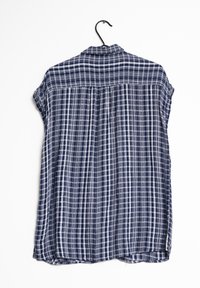 Camisa de cuadros azul marino y blanca de manga corta, con cuello abotonado y detalle de pliegues en la parte posterior, colgando en un gancho negro.