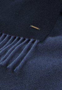 Marinblå uldscarf med frynser og en lille metallisk detalje med en gul indsats. Blød tekstur, glat overflade og ensartet farve.
