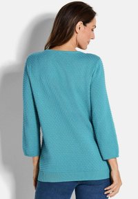 Frau mit langen braunen Haaren, die einen strukturierten türkisfarbenen Pullover mit Dreiviertelärmel und blaue Jeans trägt, von hinten gezeigt.