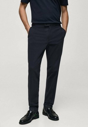 Pantaloni blu navy con vestibilità sartoriale, dotati di tasche laterali e chiusura a bottone. Abbinati a scarpe nere lucide. Tessuto liscio, design classico.