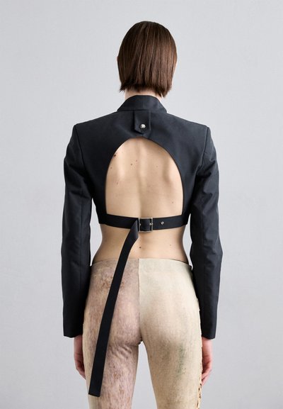 Black cropped jacket with an open back, featuring a high collar and a belt strap. The model wears patterned, skin-tone pants.
