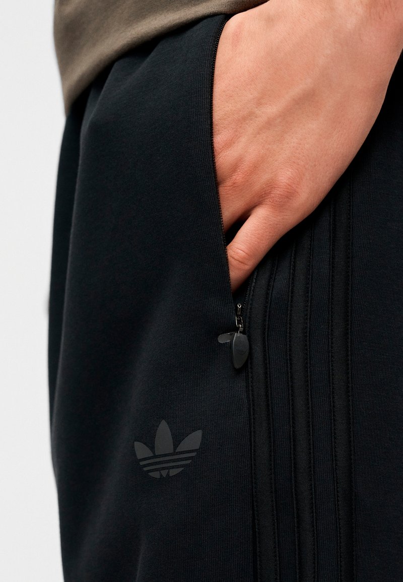 Højre hånd inde i den lynlåslomme på sorte Adidas-bukser med logo og syninger på siden.