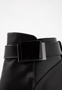 Černá kožená kotníková bota s lesklým černým čtvercovým přezkovým akcentem na pásku, s hladkým povrchem a elegantním designem.