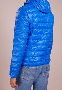 Niebieska kurtka puchowa z błyszczącym, pikowanym wykończeniem i kapturem. Posiada poziome przeszycia i dopasowany krój, zestawiona z jasnymi dżinsami.