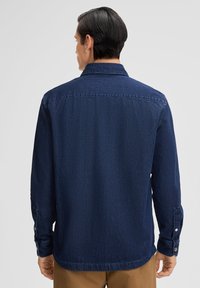 Marineblauwe denim overhemd met lange mouwen, geknoopte manchetten en een kraag. Heeft een rechte snit en een detail van een achterlijfnaad.