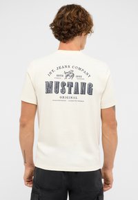 Man with curly brown hair wearing a white Mustang branded T-shirt featuring a black logo and text on the back, standing with back to camera.