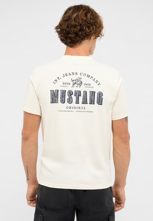 Mężczyzna z kręconymi brązowymi włosami, ubrany w białą koszulkę marki Mustang z czarnym logo i napisem z tyłu, stojący tyłem do aparatu.