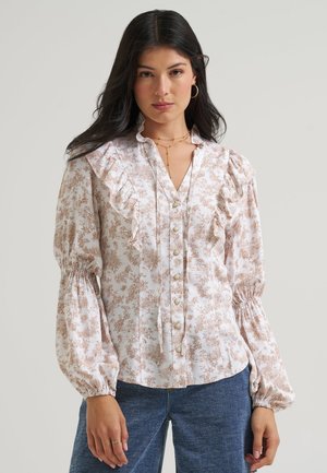 Femme portant un chemisier blanc à imprimé floral marron, détails à volants, boutons en perles et manches longues bouffantes, associé à un jean bleu.