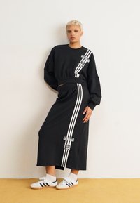 Fekete kapucnis pulóver és hozzáillő hosszú szoknya fehér csíkozással, Adidas logós folttal, klasszikus fehér sportcipővel párosítva.